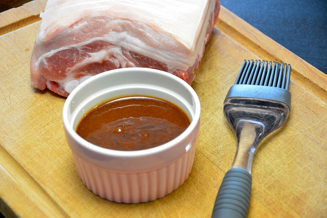 A cup of barbecue sauce, with meat behind it, and a brush next to the sauce.